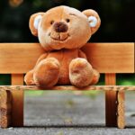 Smiling teddy bear sitting on a miniature wooden bench in a park