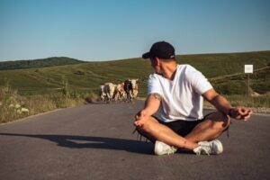 Photo of man sitting in the middle of the road with a herd of cattle coming up the road behind him