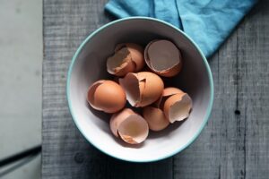 Brown egg shells in a white bowl on a wooden surface, photo taken from above.