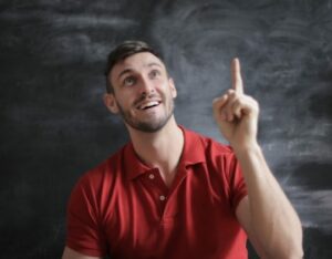 Man in a red polo shirt in front of a blackboard holding up a finger as though he's had a good idea