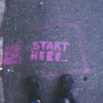 Tarmac and cobbled surface with a pair of feet and a pink "start here" message written in a square box in front of them.