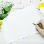 A blank piece of white paper with a person's hand hovering holding a green crayon, with white and yellow flowers beside the paper on a white table top