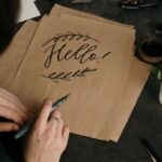 Some brown paper bags on a tabletop with a hand resting on the top holding a black pen having written 'Hello!' on the top bag, decorated with a leaf design.