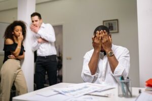 Man seated in an office covering his face with his hands while 2 colleagues look on whispering to each other.