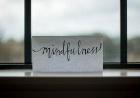A white piece of paper with the word mindfulness written on it propped on a windowsill with blurred view in the background