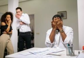 Man seated in an office covering his face with his hands while 2 colleagues look on whispering to each other.