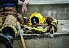 A yellow hard hat resting on a brown and yellow firefighter's suit beside a firefighter sitting taking a break